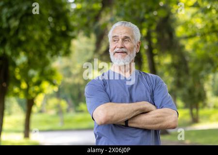 Una foto ravvicinata di un vecchio uomo dai capelli grigi in piedi in abiti casual in natura, che incrocia le braccia per terra e guarda di lato con un sorriso. Foto Stock