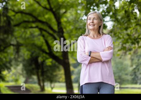 Foto di una donna anziana dai capelli grigi sorridente e rilassata in piedi in un parco con abiti casual, stringendo le mani sul petto e guardando di lato. Foto Stock