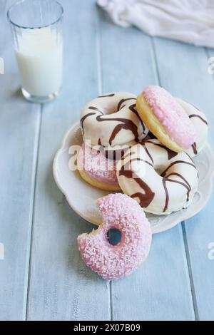 Ciambelle alla vaniglia smerigliate con vortici al cioccolato e ciambelle rosa fragola con fiocchi di cocco. Morso mancante. Bicchiere di latte sullo sfondo. Foto Stock