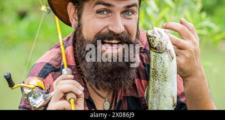 Pescatore con la canna da pesca in mano che tira il pesce pescato dal fiume. L'uomo tiene in mano una trota di pesci grossa. Pescatore e trofeo. Uomo rilassante e.. Foto Stock