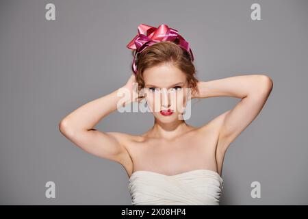 Giovane donna che trasuda pose di bellezza classiche in studio con abito bianco con fiocco rosa sulla testa. Foto Stock