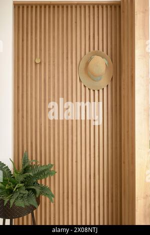 Un cappello di paglia con un nastro verde è appeso su una moderna parete a lastre di legno sopra una felce in vaso, creando un angolo tranquillo Foto Stock