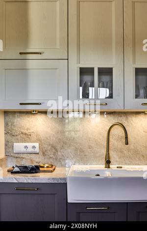 Cucina dal design moderno ed elegante con lavabo bianco della fattoria con rubinetto in ottone, adagiato su uno splendido lavabo in marmo e armadietto in legno chiaro Foto Stock