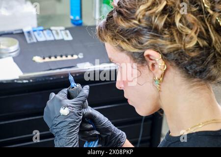 Un'artista di tatuaggi donna con guanti inchioda meticolosamente un design sulla pelle di un cliente in uno studio di tatuaggio ben attrezzato. Foto Stock