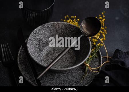 Dall'alto di un tavolo artistico caratterizzato da eleganti stoviglie nere, accentuate da vibranti fiori gialli di mimosa e da un tocco di verde. Foto Stock