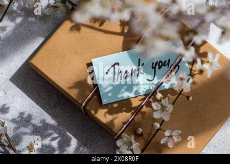 Un caloroso ringraziamento poggia su una confezione regalo di carta kraft, delicatamente circondata dai petali morbidi e dalle ombre dei ciliegi in fiore, evocando un senso di Foto Stock