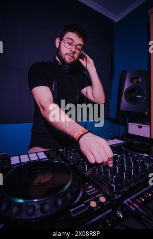 Un DJ maschile attento regola le cuffie mentre mescola la musica su una console DJ professionale in una stanza poco illuminata con illuminazione soffusa Foto Stock