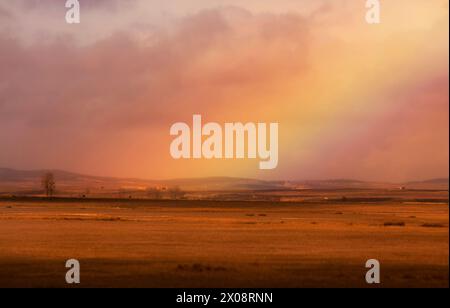 Le calde sfumature del tramonto bagnano il paesaggio come morbidi archi arcobaleno sopra un campo sereno Foto Stock