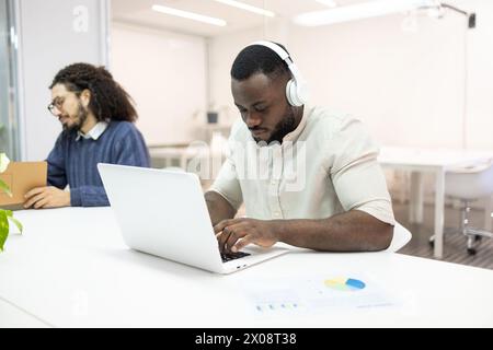 Due uomini concentrati in un ufficio moderno con un uomo che indossa le cuffie e scrive su un notebook. Foto Stock