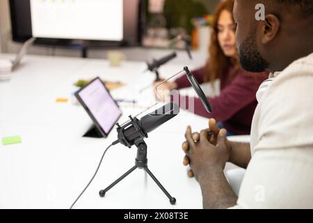 Un uomo nero ritagliato e una donna, probabilmente di etnia ispanica o mediorientale, si impegnano in una sessione di registrazione di podcast in un moderno studio, W Foto Stock