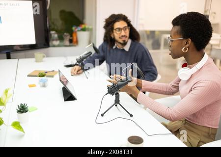 Una donna nera regola le attrezzature mentre un uomo con i capelli ricci, probabilmente di etnia mediorientale o ispanica, la guarda in un podcasting. Foto Stock