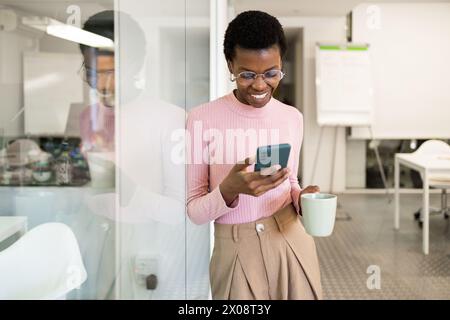Allegra donna afroamericana che guarda il suo telefono e tiene in mano una tazza da caffè, in piedi in un ambiente d'ufficio. Foto Stock