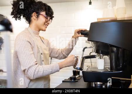 Una allegra barista ispanica con capelli ricci e bicchieri monta il latte utilizzando una macchina da caffè professionale in un ambiente luminoso. Foto Stock