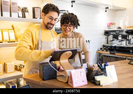 I baristi multietnici che sorridono di sesso maschile e femminile collaborano utilizzando un tablet in un ambiente accogliente e ben illuminato, mostrando lavoro di squadra e tecnologia in un piccolo spazio Foto Stock