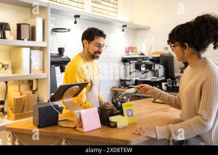 Un barista sorridente con una camicia gialla accetta il pagamento da una cliente donna utilizzando un lettore di carte presso un moderno banco di caffetteria. Foto Stock