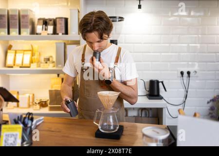 Un barista concentrato in un grembiule di tela puzza di chicchi appena macinati mentre prepara il caffè utilizzando un metodo a "pour-over" in un ambiente luminoso e alla moda. Foto Stock