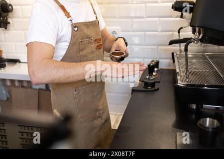 Un barista in grembiule tiene in mano un portafiltro pieno di caffè macinato, pronto per l'estrazione con una macchina per espresso in un ambiente bar. Foto Stock