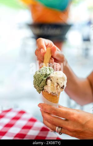Uno scatto ravvicinato che cattura la mano di una persona irriconoscibile ritagliata che tiene in mano un cono con due palline di gelato, contro un retrogroun vibrante e sfocato Foto Stock