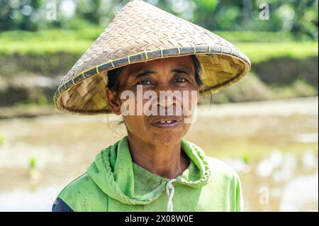 Una contadina balinese che indossa un tradizionale cappello intrecciato, in piedi di fronte a una verdeggiante risaia a Bali. La sua espressione racconta una storia di bacchetta dura Foto Stock