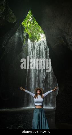 Una donna esuberante con le braccia allungate che gode della serenità di una cascata isolata a Bali, Indonesia, circondata da una vegetazione lussureggiante. Foto Stock