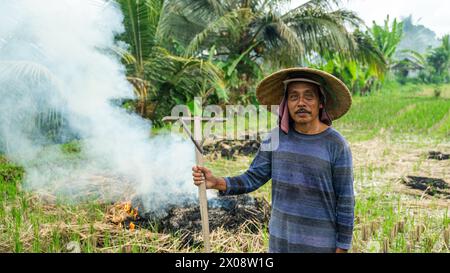 Un contadino balinese si trova in una risaia con indosso un cappello tradizionale, tenendo un rastrello con fumo proveniente dal raccolto che brucia sullo sfondo. Foto Stock