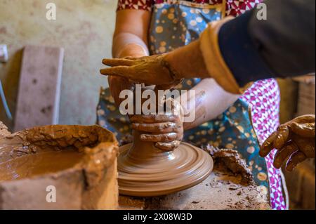 Primo piano delle mani di un artigiano corto e irriconoscibile che modellano sapientemente l'argilla bagnata in una pentola su una ruota di ceramica rotante, con spruzzi di fango intorno. Foto Stock