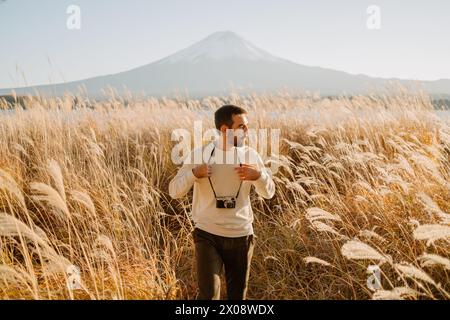 Un viaggiatore maschile con una macchina fotografica gode di vedute maestose del Monte Fuji, circondato da pampas d'oro sotto un cielo limpido Foto Stock