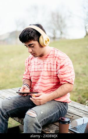 Un uomo concentrato si siede a un tavolo da picnic nel parco, ascoltando la musica con le cuffie mentre utilizza lo smartphone, con un portatile accanto a sé Foto Stock