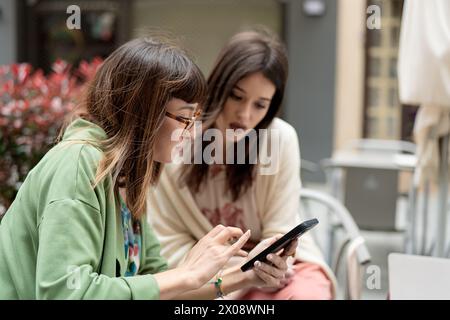 Due giovani donne, vestite con un leggero abbigliamento primaverile, condividono un momento guardando uno smartphone insieme su un patio all'aperto immerso nel verde Foto Stock