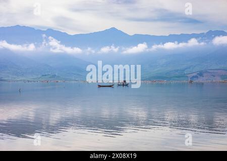 Acque tranquille di un lago nebbioso con barche tradizionali in Vietnam, sullo sfondo di montagne lussureggianti. Foto Stock