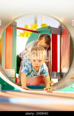Una famiglia amorevole gioca in un parco giochi dove un bambino con la sindrome di Down scorre allegramente attraverso un tunnel colorato con lo sguardo incoraggiante dei suoi genitori Foto Stock