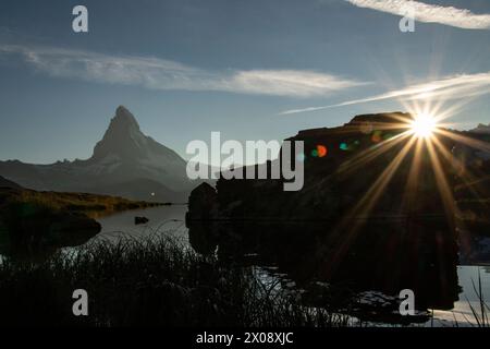 I raggi del sole esplodono su una silhouette rocciosa, con l'iconico Cervino riflesso nelle tranquille acque di Stellisee, annunciando un nuovo giorno nella SWI Foto Stock