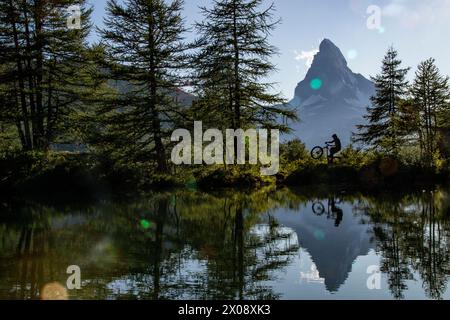 Una sorprendente silhouette di una persona in bicicletta vicino a un lago calmo, con l'iconica montagna Cervino e alberi lussureggianti che si riflettono nelle acque cristalline sottostanti Foto Stock