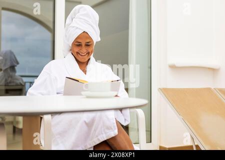 Una donna ispanica sorridente di mezza età avvolta in un accappatoio bianco e asciugamano legge un libro mentre sorseggia un caffè su un tranquillo balcone. Foto Stock