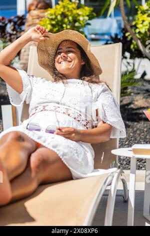 Una donna ispanica di mezza età sdraiata su una sedia a sdraio, sorridendo sotto un cappello a tegole larghe, godendosi una giornata di vacanza soleggiata, incarnando relax e contenuti Foto Stock