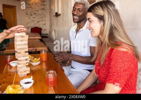 Un gruppo eterogeneo di amici condivide le Risate mentre gioca a un gioco a blocchi di legno in un bar rustico, con bevande rinfrescanti sul tavolo Foto Stock