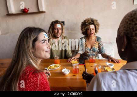 Un gruppo di amici diversi che ridono e giocano a un gioco di appiccicose note con bevande e spuntini in un rustico tavolo da bar Foto Stock