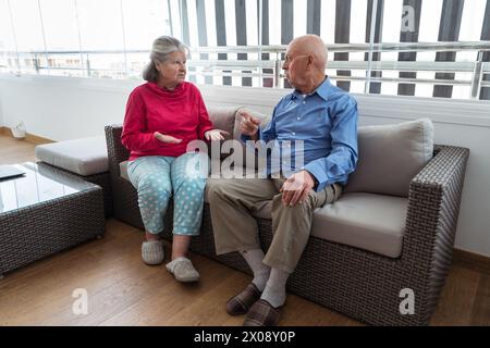 Un'anziana coppia caucasica si impegna in una conversazione ponderata mentre si siede comodamente su un divano in un soggiorno ben illuminato, suggerendo un momento di Foto Stock