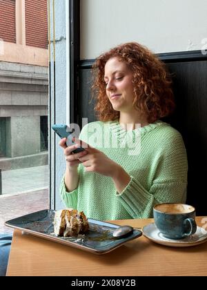 Sorridente giovane rossa che si gode una pausa caffè mentre usa il suo smartphone in un accogliente ambiente da caffè Foto Stock