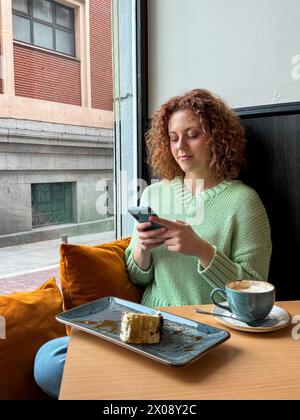 Una giovane donna rossa con i capelli ricci è fidanzata con il suo smartphone, seduta accanto a una finestra in un accogliente caffè, con un dessert a metà pasto e un cappuccino Foto Stock