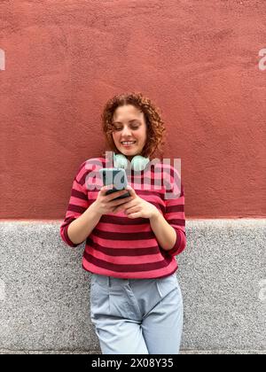 La giovane donna rossa dai capelli ricci in un maglione a righe sorride guardando il suo smartphone, con le cuffie intorno al collo, in piedi contro un rosso Foto Stock