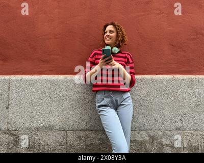 Una donna rossa sorridente con i capelli ricci si appoggia contro un muro, si impegna con il telefono e con le cuffie intorno al collo in una giornata di sole Foto Stock