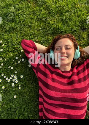 Una donna rossa gioiosa giace su un lussureggiante prato verde con margherite, indossa cuffie e una camicia a righe rosse, che incarna relax e felicità Foto Stock