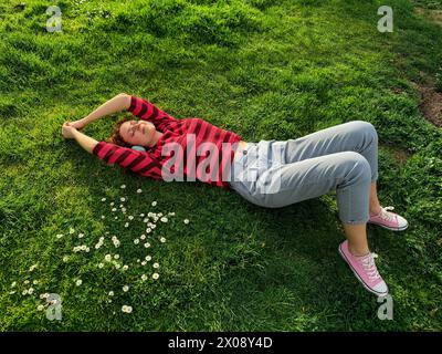 Una giovane donna rossa si trova sull'erba lussureggiante, circondata da margherite, godendosi un momento di pace nella natura Foto Stock