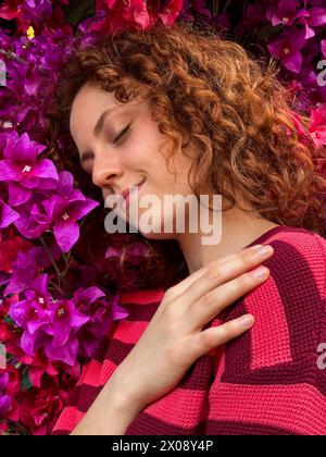 Una giovane donna rossa con capelli ricci gode un momento di pace circondato da vibranti fiori rosa Foto Stock