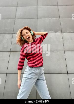 Una giovane donna rossa esuberante con capelli ricci e un maglione a righe ama la musica indossando le cuffie contro una parete grigia Foto Stock