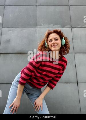 Una giovane donna rossa allegra con i capelli ricci che indossa un maglione a righe rosse e nere si appoggia contro una parete grigia mentre ascolta la musica sul suo tè Foto Stock