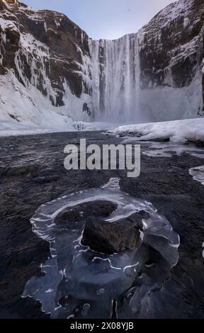 Una foto impressionante che cattura la bellezza grezza di una cascata ghiacciata in Islanda, circondata da scogliere ghiacciate e da un tranquillo letto di fiume con intricati ghiacci Foto Stock