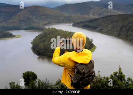 La vista posteriore di un uomo con giacca gialla e berretto si erge su un punto panoramico sopraelevato, scattando una foto di un fiume tortuoso in un lussureggiante paesaggio montano. Foto Stock