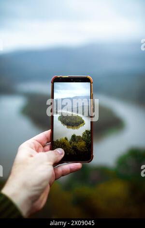 Una mano regge uno smartphone, incorniciando un pittoresco fiume circondato da alberi sullo sfondo nebbioso. Foto Stock